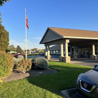 A building with a flagpole displaying the American flag, surrounded by greenery and parked cars on a clear day.