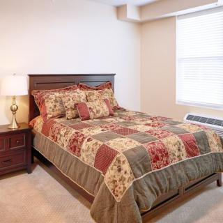 A cozy bedroom featuring a wooden bed with a patchwork quilt, a nightstand with a lamp, and natural light streaming through the window.