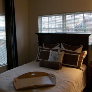 A cozy bedroom with a neatly made bed and decorative pillows, illuminated by natural light from the windows.