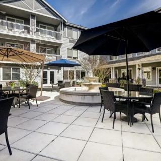 A modern outdoor courtyard featuring patio furniture, umbrellas, and a central fountain in front of a multi-story building.