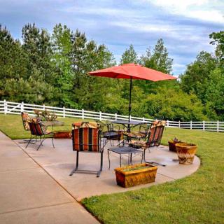 A serene backyard patio with outdoor furniture and a red umbrella, surrounded by greenery and a white fence.