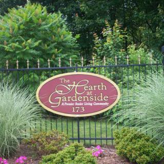 A decorative garden sign labeled 'The Hearth at Gardenside' mounted on a fence surrounded by lush greenery.
