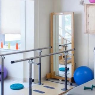A bright physical therapy room with various exercise equipment, including parallel bars, a balance ball, and a mirror.