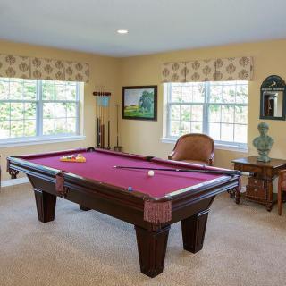 A well-lit game room featuring a billiards table with magenta felt, surrounded by windows and decorated with artwork and furniture.