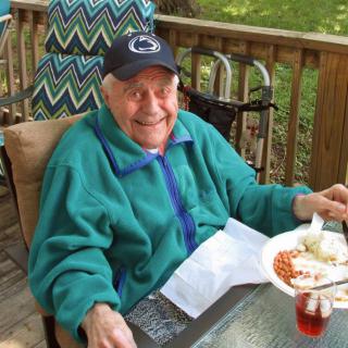 An elderly man enjoys a meal outdoors, smiling while wearing a cap and green jacket.