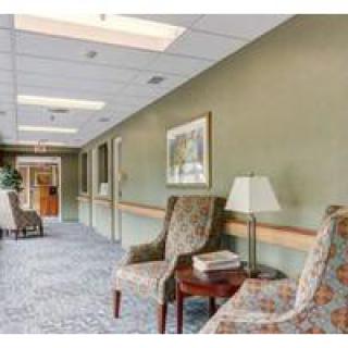 A hallway with upholstered chairs and a table topped with a lamp, featuring carpeted flooring and wall decor.