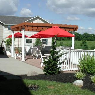 A sunny patio area with a pergola, red umbrellas, and outdoor seating next to a house.