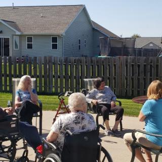A group of elderly people sitting outdoors, some in wheelchairs, enjoying a sunny day in a garden area.