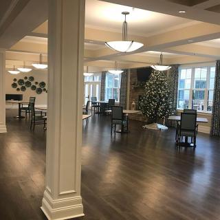 A spacious dining area with elegant chandeliers, hardwood flooring, and a decorated tree by large windows.