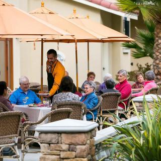 A group of seniors enjoying a meal outdoors at a restaurant patio, attended by a waiter.