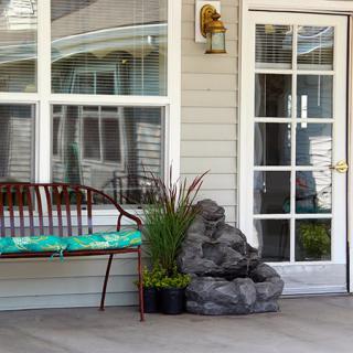 A cozy porch setup with a bench, cushion, plants, and a decorative rock next to a glass door.
