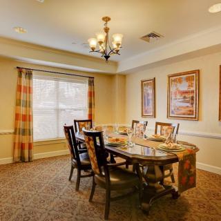A cozy dining room with a chandelier, wooden table and chairs, framed art on the walls, and a large window with curtains.
