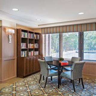 A cozy reading area featuring a wooden bookshelf filled with books, a round table surrounded by chairs, and plenty of natural light from large windows.