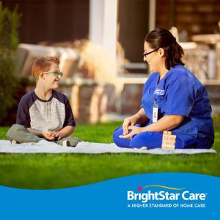 A caregiver and a child sit on a blanket outdoors, engaging in conversation.