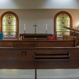 An interior view of a church showcasing wooden pews and a simple altar with stained glass windows.
