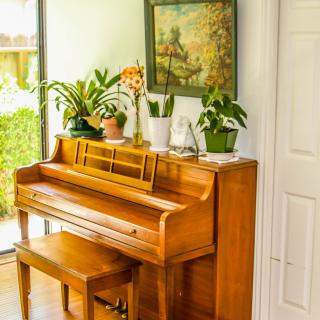 A cozy interior featuring a wooden piano adorned with potted plants and artwork, illuminated by natural light from a nearby window.