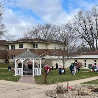 A community gathering outdoors near a gazebo with buildings in the background and trees indicating an autumn setting.