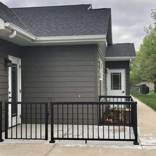 A modern house exterior with a small patio area enclosed by black railings and adjacent sidewalk.