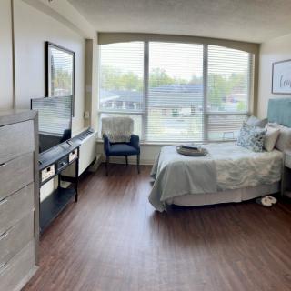 A modern bedroom with large windows allowing natural light, featuring a bed, dresser, and TV setup.