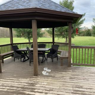 A wooden gazebo with patio furniture on a deck overlooking a grassy field and trees.