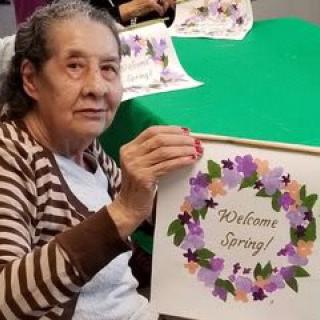 Elderly woman holding a 'Welcome Spring' craft with floral design.