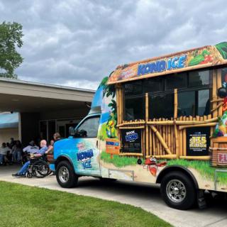 A colorful ice cream truck parked outside a building on a cloudy day, with people gathered nearby.