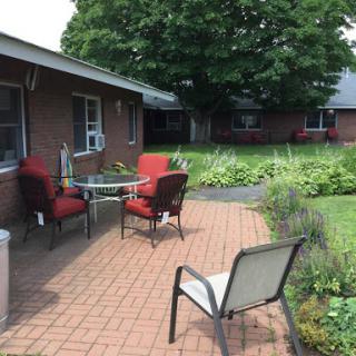 A cozy outdoor patio area with red chairs and a table, surrounded by greenery.