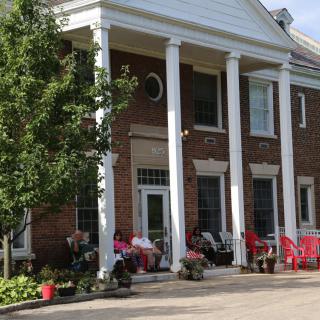 A brick building with large white columns and a porch decorated with chairs and plants.