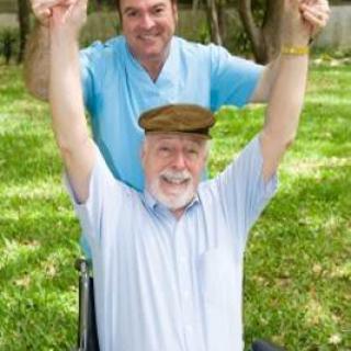 An elderly man in a wheelchair raising his arms joyfully, accompanied by a caregiver outdoors.