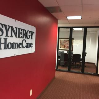 A corporate office environment with a SYNERGY HomeCare sign on a red wall, leading to a conference room.