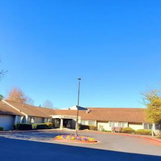 A single-story building with a tan roof, surrounded by trees and a parking lot, under a clear blue sky.
