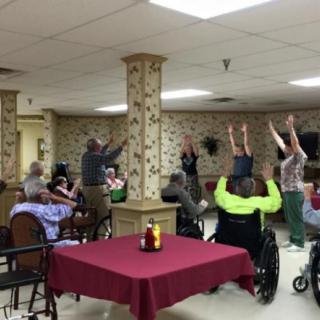 A group of elderly individuals participating in a seated exercise session, some using wheelchairs, in a communal setting.