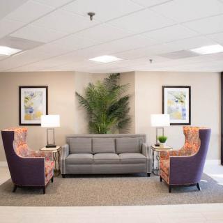 A cozy lobby area featuring a grey sofa and patterned chairs with lamps on side tables, framed artwork on the walls, and a large indoor plant.