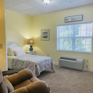 A cozy bedroom with a bed, lamp, and comfortable chair, featuring a large window and carpeted floor.