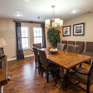 A cozy dining room with a large wooden table, surrounded by chairs and illuminated by a chandelier. The room features artwork on the walls, hardwood flooring, and natural light pouring in through double windows adorned with curtains.