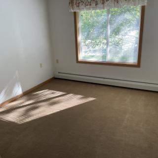 An empty room with a large window allowing sunlight to illuminate the carpeted floor.