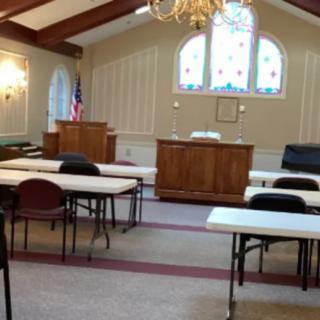 Interior of a church meeting room with tables and chairs arranged, featuring a pulpit and stained glass windows.