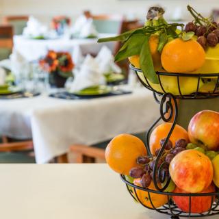 A fruit basket with oranges, peaches, and grapes sits in the foreground, while a neatly set dining table is blurred in the background.