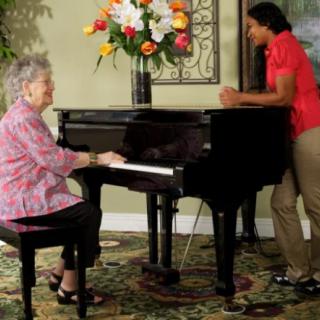 An elderly woman plays the piano while a caregiver listens attentively.