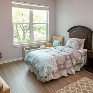 A cozy bedroom with a single bed, wooden floor, and ample natural light coming through a large window.