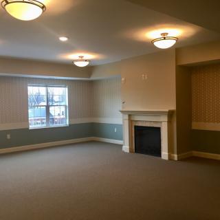 A spacious living room featuring a fireplace, carpeted floor, and large window with ceiling lights.