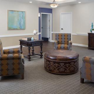 A cozy lounge area featuring comfortable seating with patterned chairs and a central round ottoman, complemented by artwork and decorative pieces.