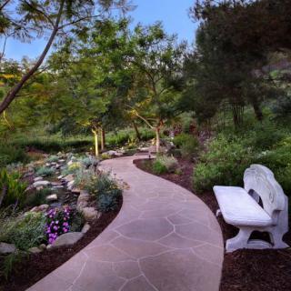 A peaceful garden pathway with a stone bench surrounded by trees and colorful flowers.