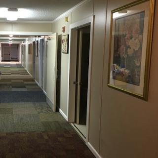 A dimly lit hotel hallway featuring carpeted floors, doors leading to rooms, and a framed artwork on the wall.