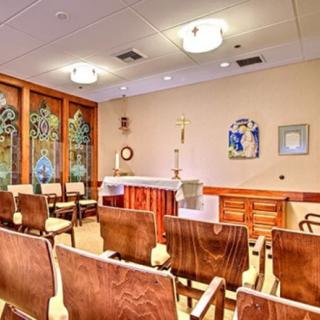 A serene chapel interior with rows of chairs facing an altar, adorned with decorative windows and soft lighting.