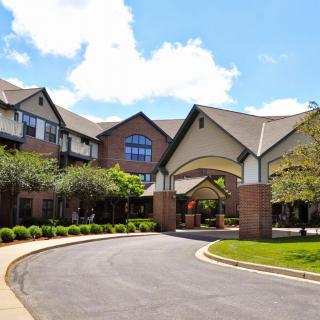 A large residential building with a curved driveway and manicured landscaping under a partly cloudy sky.