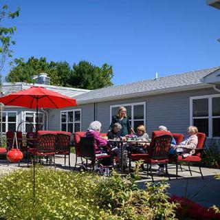 A group of elderly people socializing on an outdoor patio with red chairs and a large umbrella under a clear blue sky.