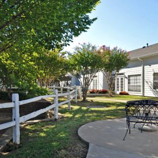 A peaceful backyard garden featuring a white fence, trees, and a patio area with seating.