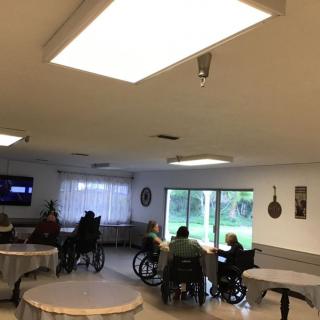 A nursing home common area with seniors in wheelchairs watching television.