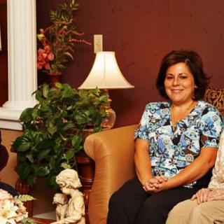 A group of people sitting in a warmly decorated room with a lamp and plant in the background.